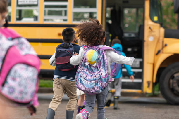 Diverse group of happy children getting on school bus A multi-ethnic group of elementary age children are getting on a school bus. The kids' backs are to the camera. They are running towards the school bus which is parked with its door open. It's a rainy day and the kids are wearing jackets, rain boots and backpacks. school children stock pictures, royalty-free photos & images