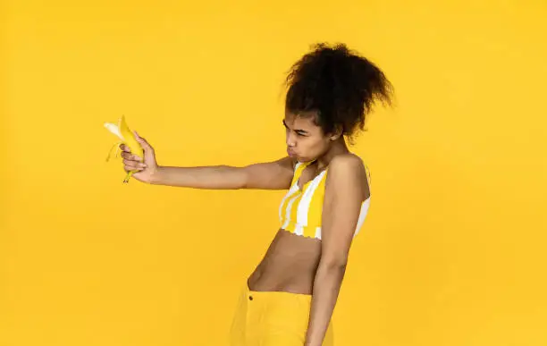 Photo of Funny dangerous african american teen girl pretending holding banana gun pistol looking aiming, silly cool black young ethnic woman make shooting gesture isolated on summer yellow studio background