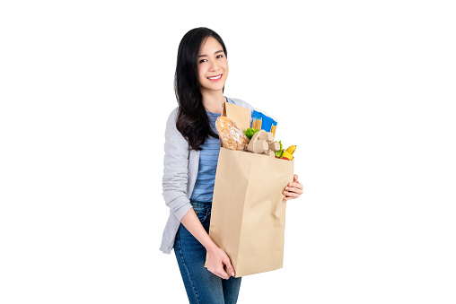 Beautiful smiling Asian woman holding paper shopping bag full of vegetables and groceries studio shot isolated on white background