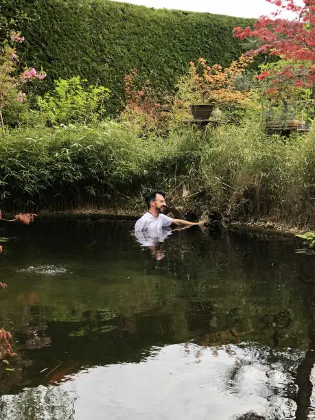 Photo of Image of man standing in large deep koi pond in winter / spring cleaning leaves, dirty cold water and pond weed blanketweed, checking pump and filter pipes in landscaped oriental garden, Japanese maples, koi carp fish goldfish water feature brick edging