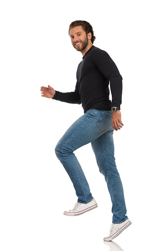Smiling young man in jeans, sneakers and black jersey is going up the stairs, smiling and looking at camera. Full length studio shot isolated on white.