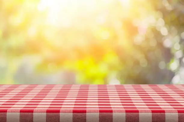 Empty table covered with red and white checkered tablecloth with defocused lush foliage at background. Ideal for product display on top of the table. Predominant color are yellow and red. DSRL studio photo taken with Canon EOS 5D Mk II and Canon EF 100mm f/2.8L Macro IS USM.