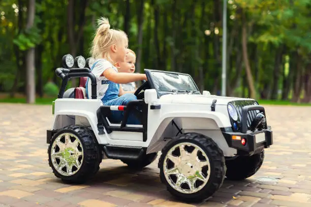 Photo of Two cute adorable blond sibings children having fun riding electric toy suv car in city park. Brother and sister enjoy playing and driving vehicle on city street outdoor. Happy childhood concept