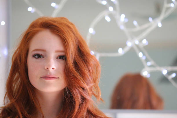 imagen de adolescente pelirrojo adolescente 14 / 15 años mirando la cámara estaba delante del espejo de la pared iluminado por luces de hadas, pelo de jengibre estilizado en ondas, maquillaje natural, piel pálida y pecas - 13 14 years teenager teenagers only female fotografías e imágenes de stock