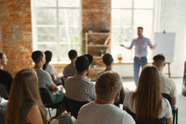 Male speaker giving presentation in hall at university workshop Male speaker giving presentation in hall at university workshop. Audience or conference hall. Rear view of unrecognized participants in audience. Scientific conference event, training. Education concept. education building stock pictures, royalty-free photos & images