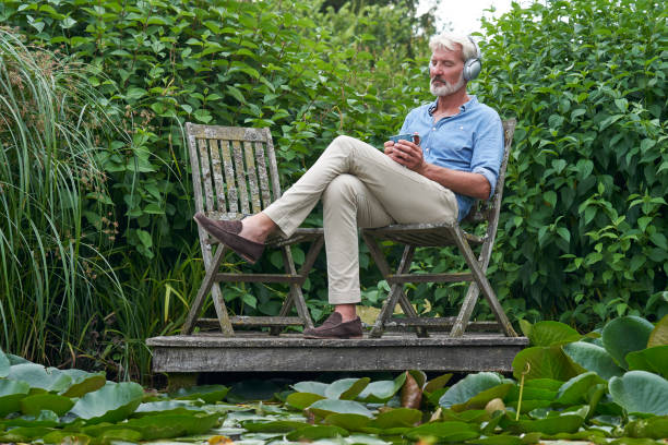 homme mûr détendant dans le jardin écoutant la musique sur des écouteurs sans fil sur la jetée par lac - the thinker audio photos et images de collection