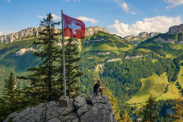 Tourist enjoys spectacular views over Appenzell Alps under Swiss flag sitting on the Ebenalp mountain in the Swiss Alps of Switzerland.