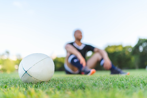 A rugby ball is put on grass field while a rugby player is sitting on grass in the background. Focus is on the rugby ball.