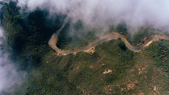 Aerial scenery view of road pass in rocky mountains covered with fog. Bird's eye view through clouds of serpentine asphalt road twists in mountainous terrain on steep slopes
