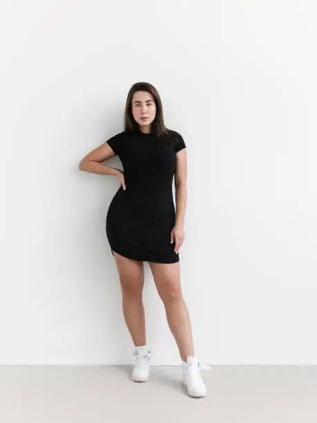 Portrait of young beautiful woman with natural make-up wearing black mini dress white sneakers looks fresh and lovely Perfect glowing skin Plus size Fashion model studio portrait on white background Full length