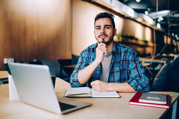 istock Pensive hipster guy looking at camera concentrated on doing homework task in library sitting with laptop computer,creative male author pondering on writing article for publication in network profile 1167037810