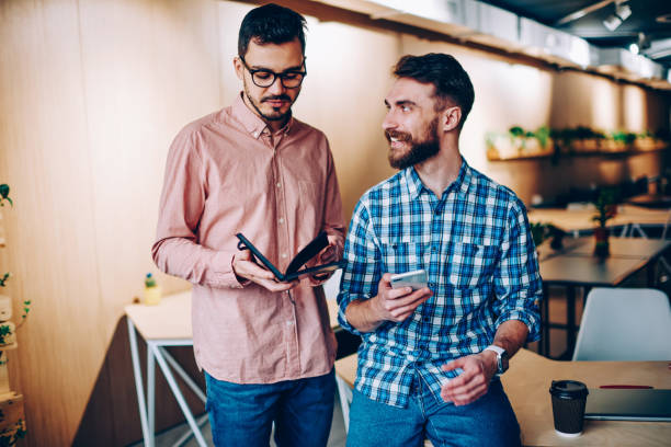istock Cheerful hipster guy looking at his colleague smiling communication in office about planning job using smartphone , man holding notepad with information discussing with coworker for cooperating 1167038399