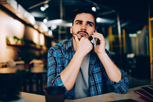 istock Contemplative hipster guy thinking aver plans for meeting while talking on telephone with friend in cafe, pensive young man waiting for connection with customer support line on mobile phone 1167038460