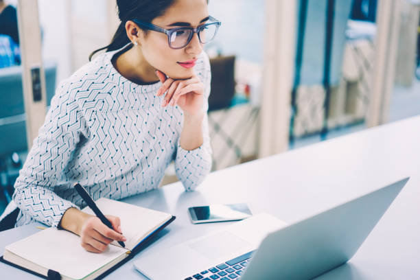 istock Pensive female administrative assistant checking report on laptop reading information and noting data,skilled woman watching online video online pondering on creating publication writing in notepad 1167039914