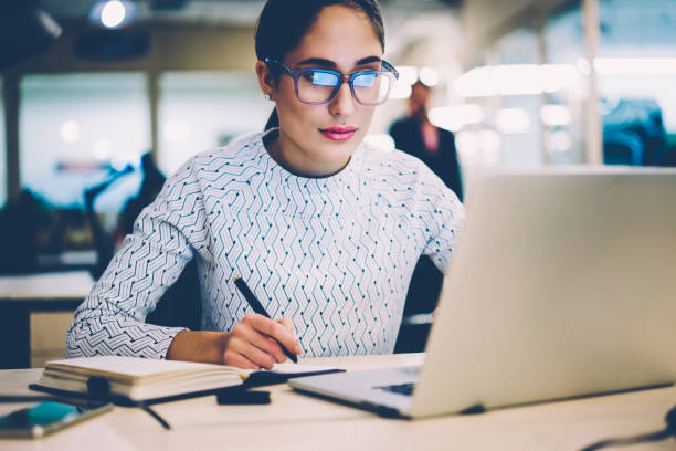 istock Serious female student in eyewear writing in netbook information from web page browsed on laptop computer,woman creating coursework project sitting at desktop with netbook for making research 1167040647