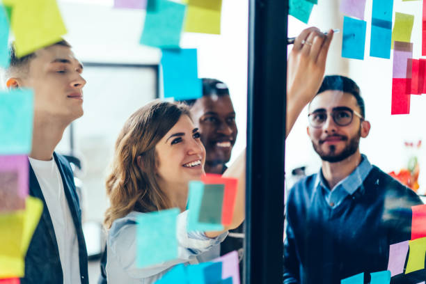 istock Diverse team of positive young people laughing while working together during brainstorming and standing behind glass wall with sticky colorful papers.Cheerful students learning words from stickers 1167050265