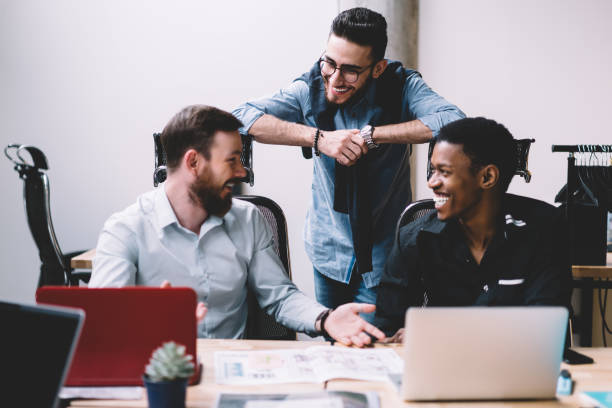 istock Multicultural young men colleagues in formal wear communicating with each other and laughing during discussion sitting at meeting table with laptops.Diverse employees friends talking about funny ideas 1167050414