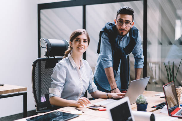 istock Portrait of male and female colleagues looking at camera while teamworking on developing internet website at modern laptop computer.Two designers collaborating on creating graphic on netbook in office 1167050505