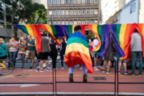 Blurred street vendor (on his back) selling LGBT flags with rainbow symbol, during pride parade on "Avenida Paulista" in sunny day. Abstract concept. Unidentified man on his back selling colorful LGBT flags hanging on the street for sale during gay parade in Sao Paulo. sao paulo gay pride parade stock pictures, royalty-free photos & images
