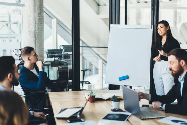istock Smart female coach conducting business training for executive managers dressed in formal wear in office.Creative young woman standing near flip chart and telling about project during conference 1167057928