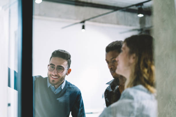 istock Positive multicultural team of young people in formal ear collaborating on common task standing behind glass wall office.Cheerful diverse employees cooperating on solution having brainstorming meeting 1167058000