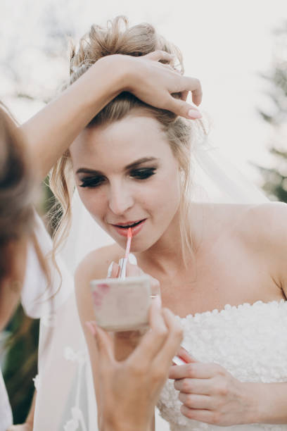 istock Stylish bride correcting her makeup, applying lipstick at wedding reception. Bridesmaid helping bride with make up, holding little mirror at wedding party 1167062202