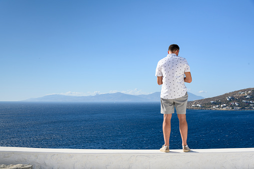 A man in a white shirt with short sleeves in shorts is standing on a background of blue sky and sea.
