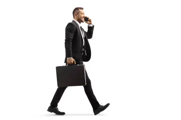 Full length profile shot of a businessman walking and talking on a mobile phone isolated on white background