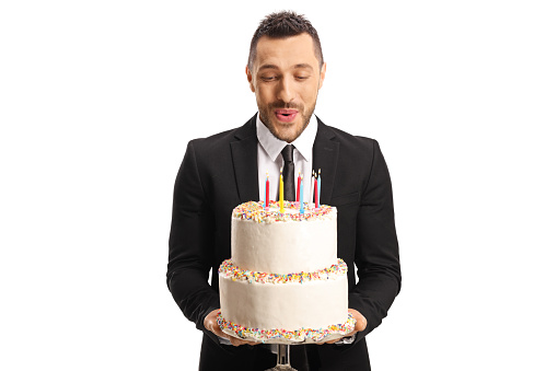 Young man in a suit holding a birthday cake and blowing candles isolated on white background
