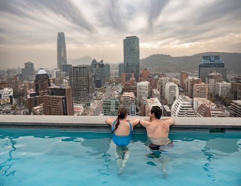 Latin american couple enjoying the swimming pool leaning at the edge of the pool looking at a beautiful city view from the rooftop of a luxury hotel