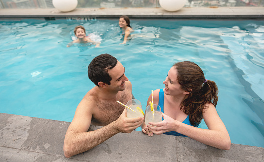 Happy adult couple having fun in the pool enjoying drinks and toasting while kids swim at background smiling - Lifestyles
