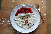 Image of children's Xmas Christmas Santa pancakes recipe / Father Christmas pancake breakfast with whipped spray cream beard, marshmallow and red strawberries for hat and nose, raisin eyes, chocolate icing mouth, Santa Claus food, breakfast for children