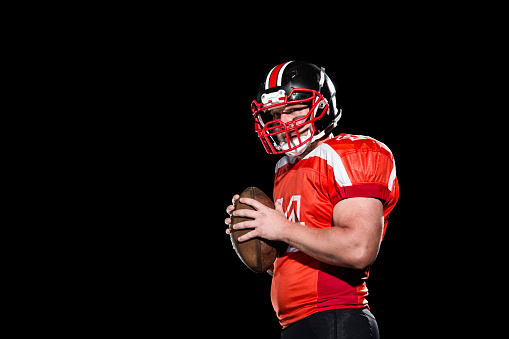 American football player in white dress and helmet holding a ball and looking where to throw the ball. Isolated on black background.