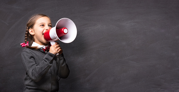 Child Girl Student Shouting Through Megaphone on Blackboard Backdrop with Available Copy Space. Back to School Concept.