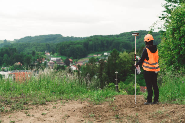 Woman Land surveyor measuring the land with topographic equipment GPS for land survey high precision measurement. Surveyor engineer in protective wear and red helmet using geodetic equipment. Female geodesist in orange vest measuring the land with total station.