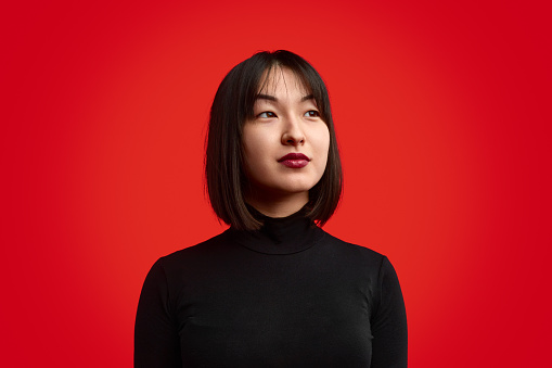 Young Japanese female in stylish black sweater looking away and thinking against vivid red background
