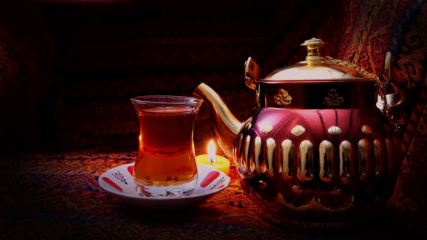 hot turkish glass of red tea with artistic teapot. old arabic teapot with smoke over dark background. arabic objects over warm fabric. middle east elements. ramadan background. arabian nights. - arabian nights imagens e fotografias de stock