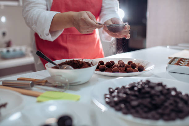 un chinois chinois asiatique mi adulte femme dessert chef époussetage et revêtement truffes au chocolat dans la cuisine domestique - saupoudreuse à farine photos et images de collection