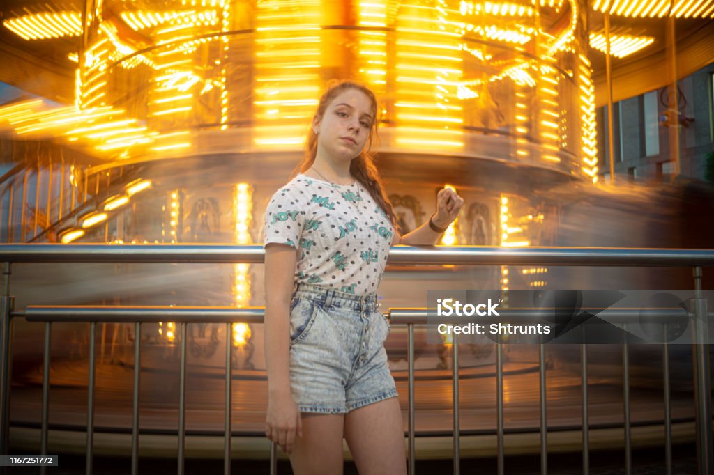 Teenage girl stands close to a spinning carousel Girl is looking down at camera. Photo was taken at evening so carousel is shining with bright orange lights. Place on this photo is central square in Dnipro city. The photo is defocused a little and contains blurred motion. It is because it was taken with long exposure and from low angle. 12-13 Years Stock Photo