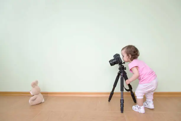 Cute funny beautiful baby in a pink T-shirt photographs her bunny. The baby stands and holds a DSLR camera on a tripod against the background of a green wall. Concept novice beginner photographer, first steps.