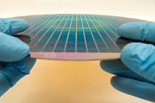 This is a patterned 300 mm silicon wafers on a white background.  Electronic circuit designs have been built onto the wafers using micromaching including photolithography.  The different colors on the wafer come from the different materials on its surface.  A person is holding the wafer with two gloved hands.