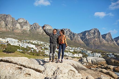 Young South African couple standing on rock formation to create selfie at foot of Twelve Apostles mountain range in Cape Town.