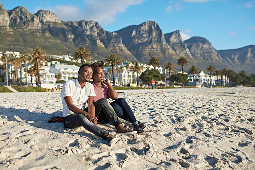 Young South African couple sitting on a white sand beach at the foot of the Twelve Apostles mountain range in Cape Town.