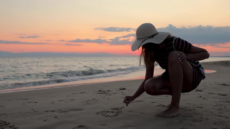 Teenage girl with sun hat on drawing love message in sand during beautiful sunset