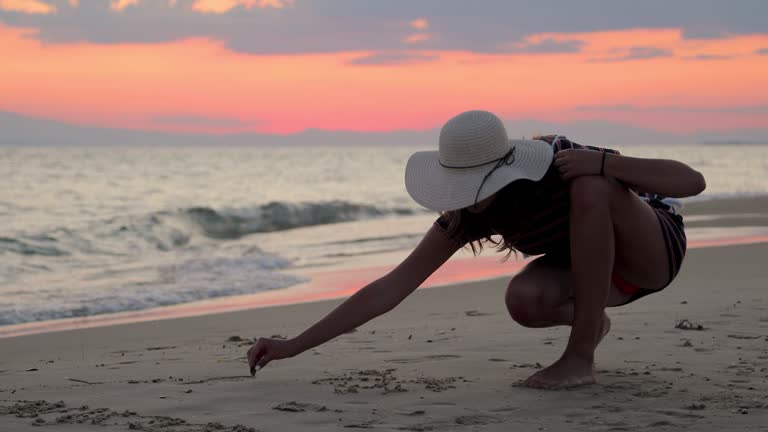 Teenage girl with sun hat on drawing love message in sand during beautiful sunset