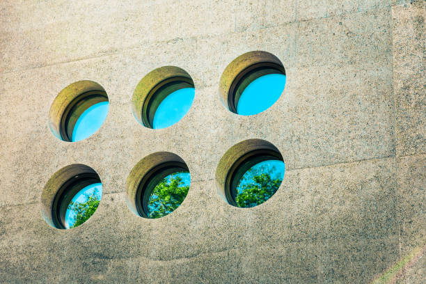 Color image depicting abstract exterior architecture in the city. The image is full of leading lines and strong geometric shapes and angles of the facade of a modern glass and steel apartment building in the city of London, UK. There are six circular windows reflecting a blue sky and lush green trees. Room for copy space.
