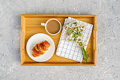 Cup of coffee with milk, freshly baked croissant, checkered napkin and camomile flowers on wooden tray on gray stone table. Concept Good morning Top view