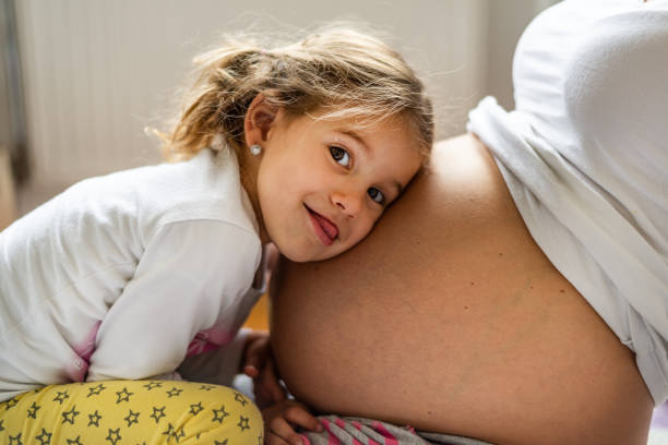 Close -up of a beautiful girl hugging mother’s pregnant belly with a smile.