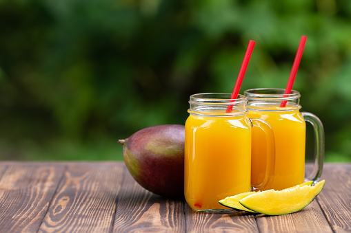 fresh mango juice in two glass mason jars with ripe fruit on wooden table outdoors. Summer refreshing drink