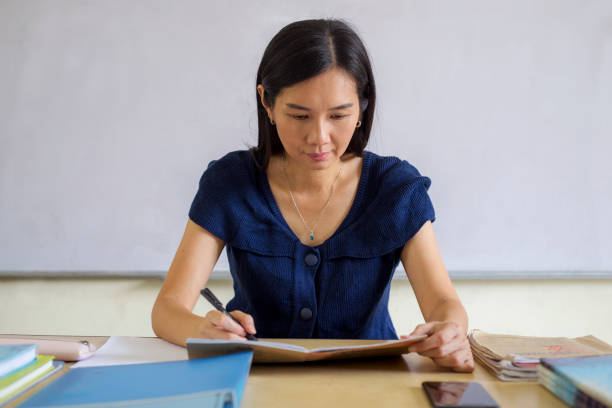 Serious female Asian teacher at her desk marking student work stock photo